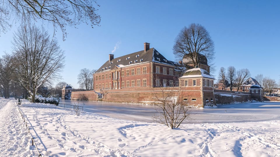 A snow-covered landscape with a historic brick building, a frozen pond, and a church spire in the background.