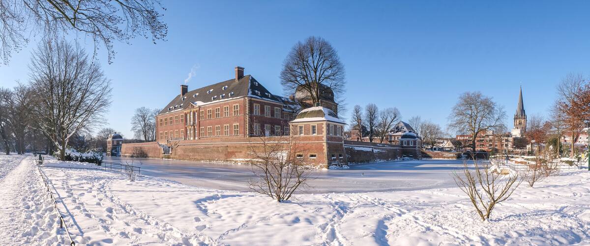 A snow-covered landscape with a historic brick building, a frozen pond, and a church spire in the background.