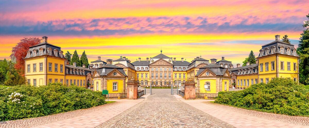 A grand yellow building with a symmetrical facade, a cobblestone pathway leading to the entrance, and a backdrop of a sunset sky.