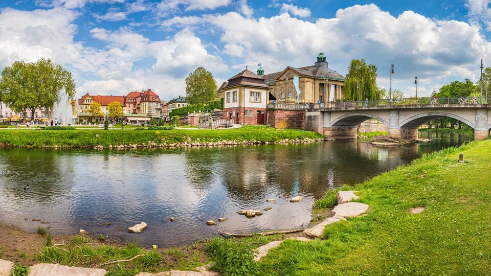 A river with a stone bridge, a fountain, and buildings in the background.