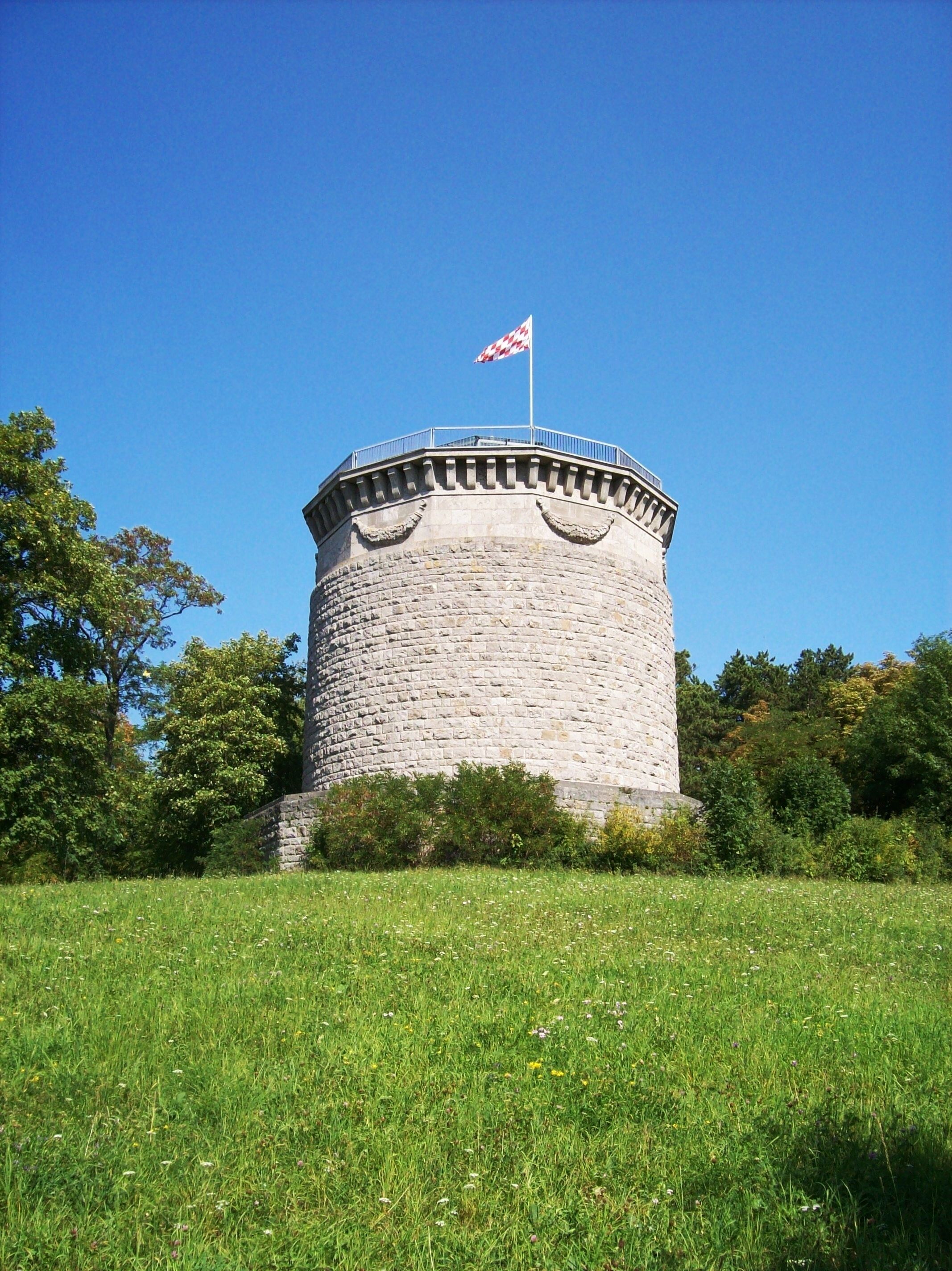 A stone tower with a flag on top, surrounded by grass and trees.