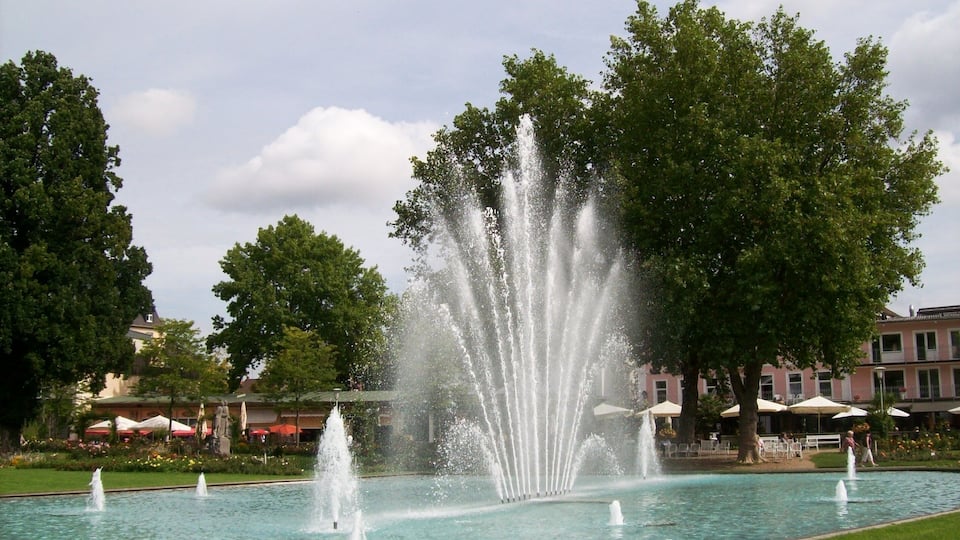 A fountain in a park with trees and buildings in the background.