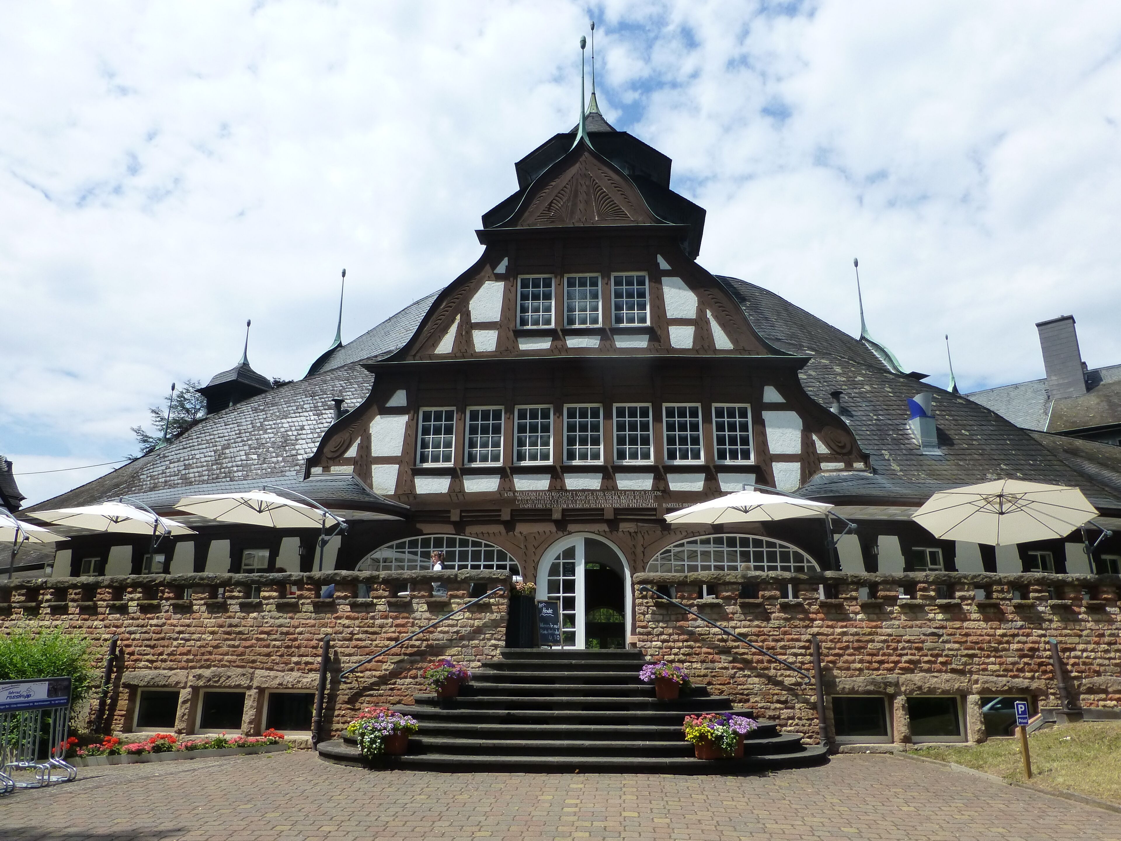 A traditional half-timbered building with a steeply pitched roof, a central entrance, and flower pots on the steps.