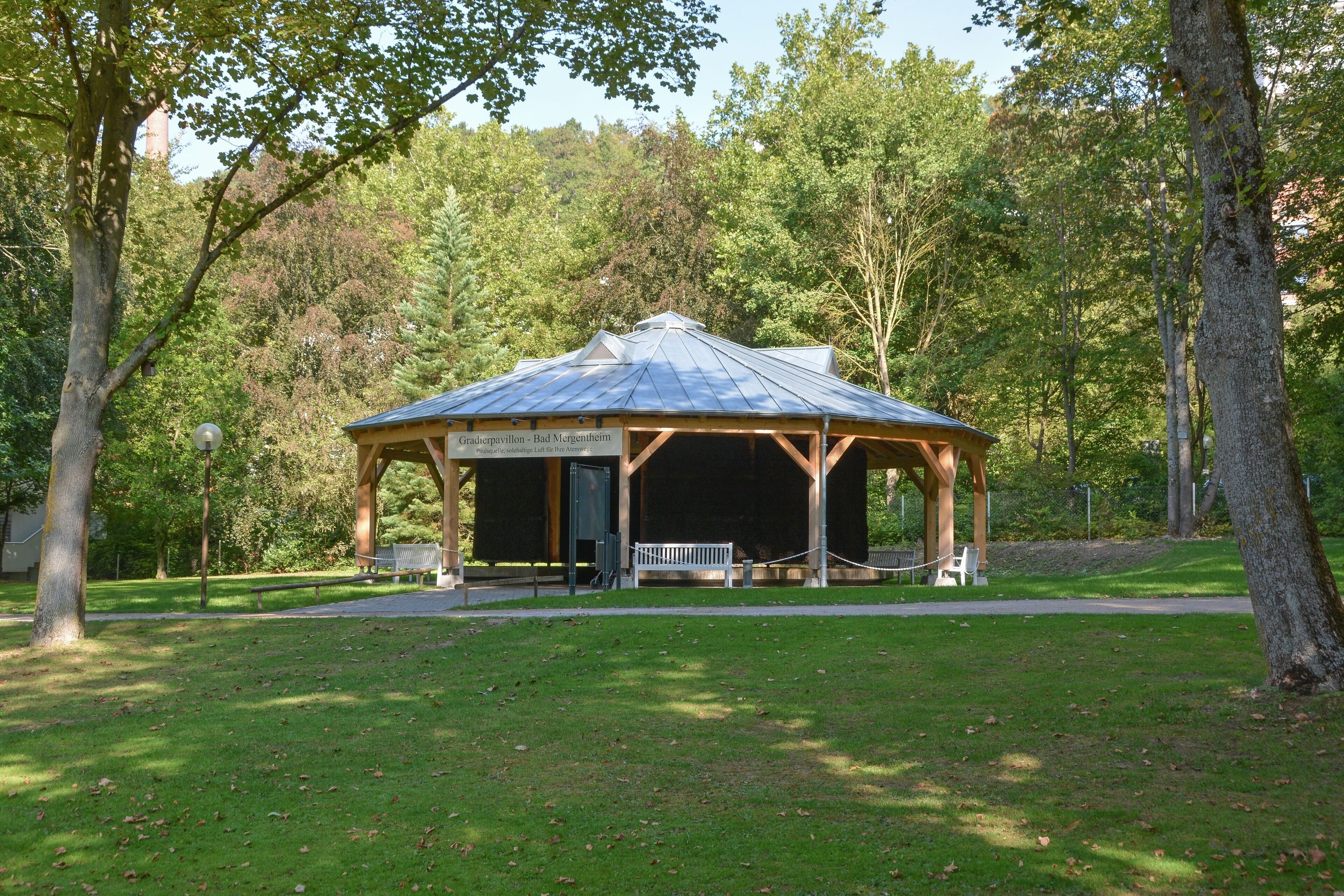 Un gazebo in un parco con tetto in metallo e travi di legno, circondato da alberi e prato.
