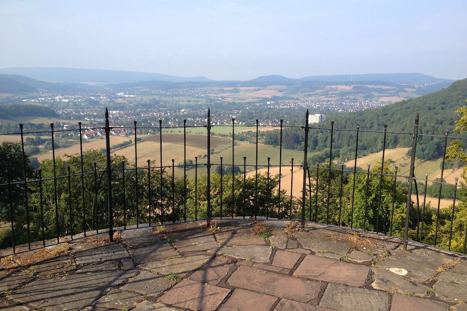 A stone-paved viewing platform with a metal fence overlooking a rural landscape with fields and scattered buildings.