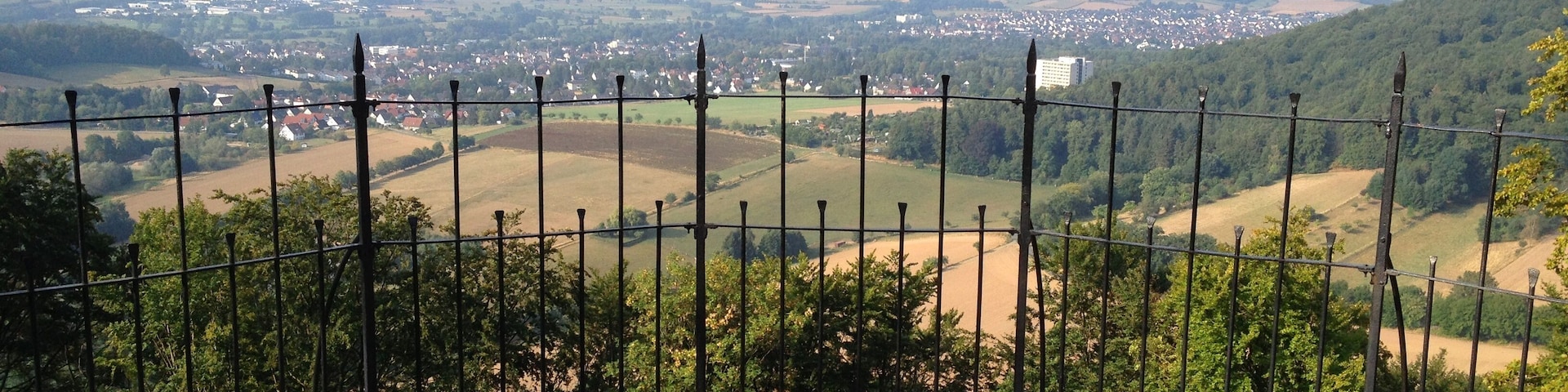 Eine Aussichtsplattform mit Steinpflasterung und Metallzaun, die auf eine ländliche Landschaft mit Feldern und verstreuten Gebäuden blickt.