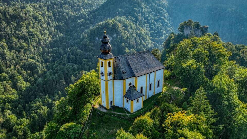 A church with a bell tower on a hill surrounded by dense forest and mountains.
