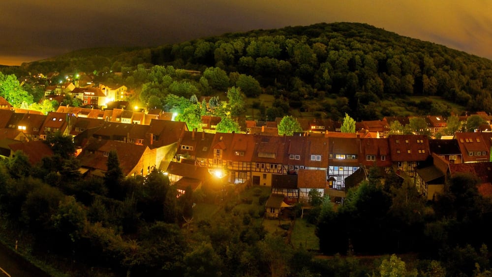 Ein Dorf bei Nacht mit beleuchteten Häusern und einem bewaldeten Hügel im Hintergrund.