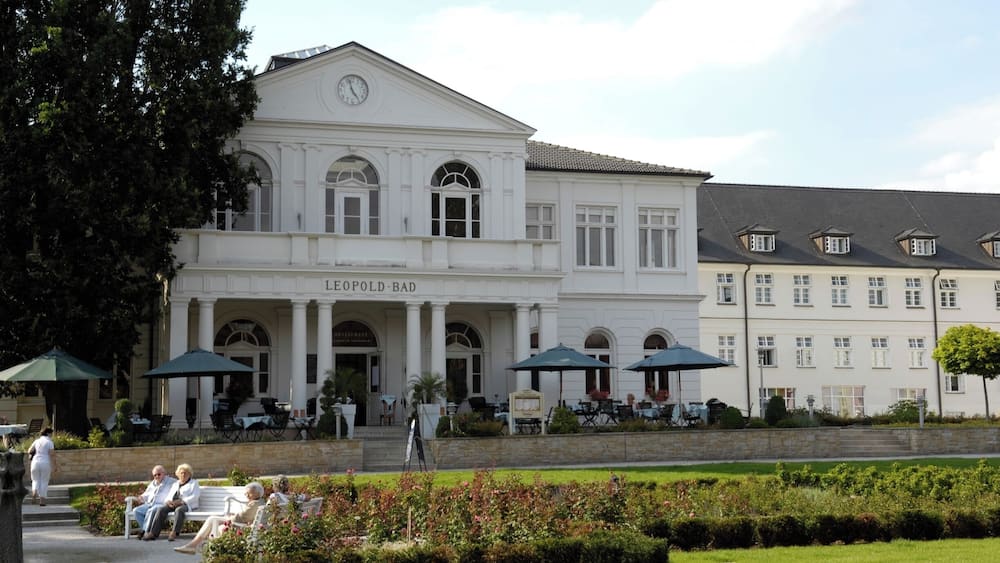A historic building with columns and a clock, surrounded by a garden and outdoor seating.