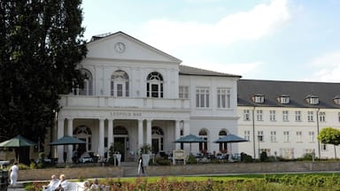 Un edificio histórico con columnas y un reloj, rodeado de un jardín y mobiliario de exterior.