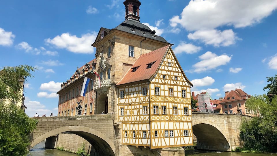Un bâtiment historique à colombages, doté d’une tour horlogée, d’un pont de pierre et bordé par une rivière.