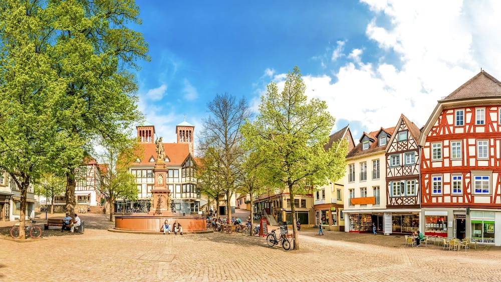 A cobblestone square with traditional half-timbered buildings, a central fountain, and people sitting on benches.