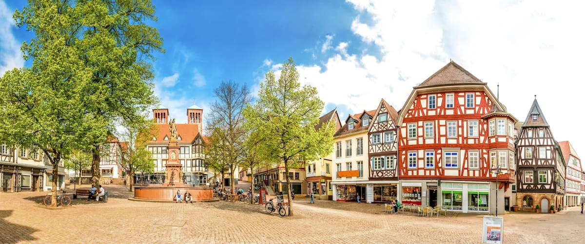 A cobblestone square with traditional half-timbered buildings, a central fountain, and people sitting on benches.