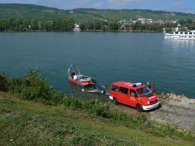 A lake with a red boat and a red vehicle near the shore, surrounded by greenery and a distant town.