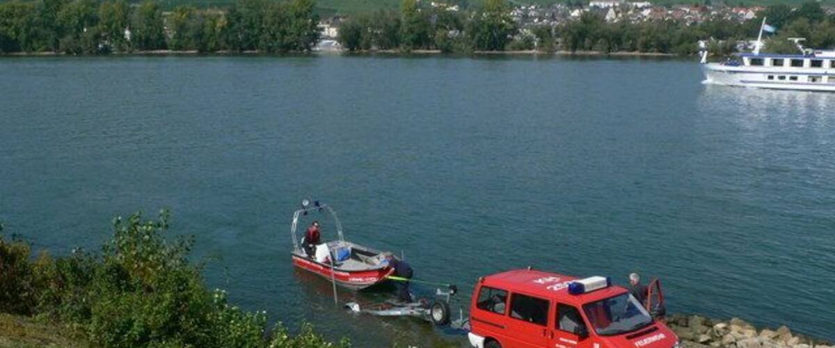 A lake with a red boat and a red vehicle near the shore, surrounded by greenery and a distant town.