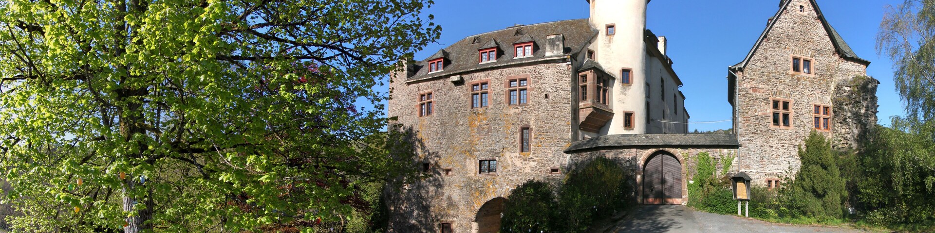 Un castillo de piedra histórico con una torre redonda, rodeado de vegetación exuberante y un cielo azul despejado.