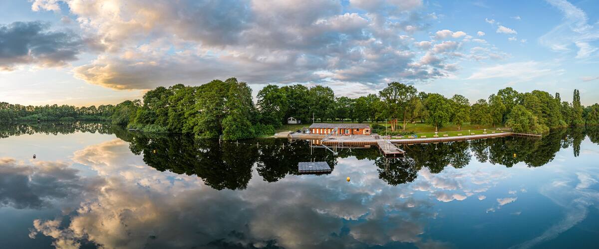 A serene lake with a dock and a small building, surrounded by dense trees and a clear sky.