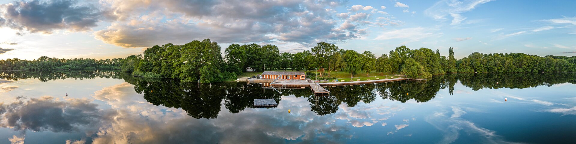 A serene lake with a dock and a small building, surrounded by dense trees and a clear sky.