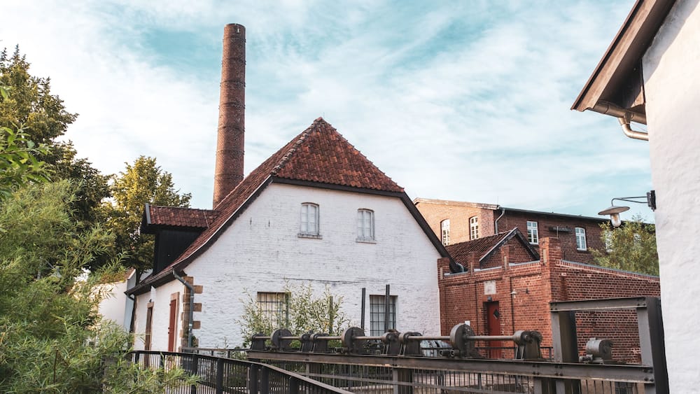 A white building with a red roof, a tall chimney, and a metal fence in front.