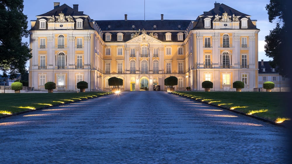 A grand, illuminated building with symmetrical architecture, a cobblestone pathway leading to the entrance, and well-manicured lawns.