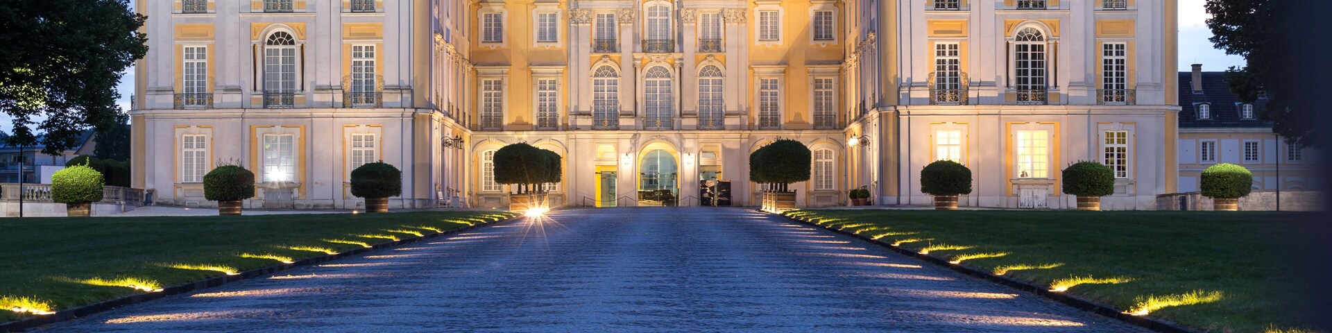 A grand, illuminated building with symmetrical architecture, a cobblestone pathway leading to the entrance, and well-manicured lawns.