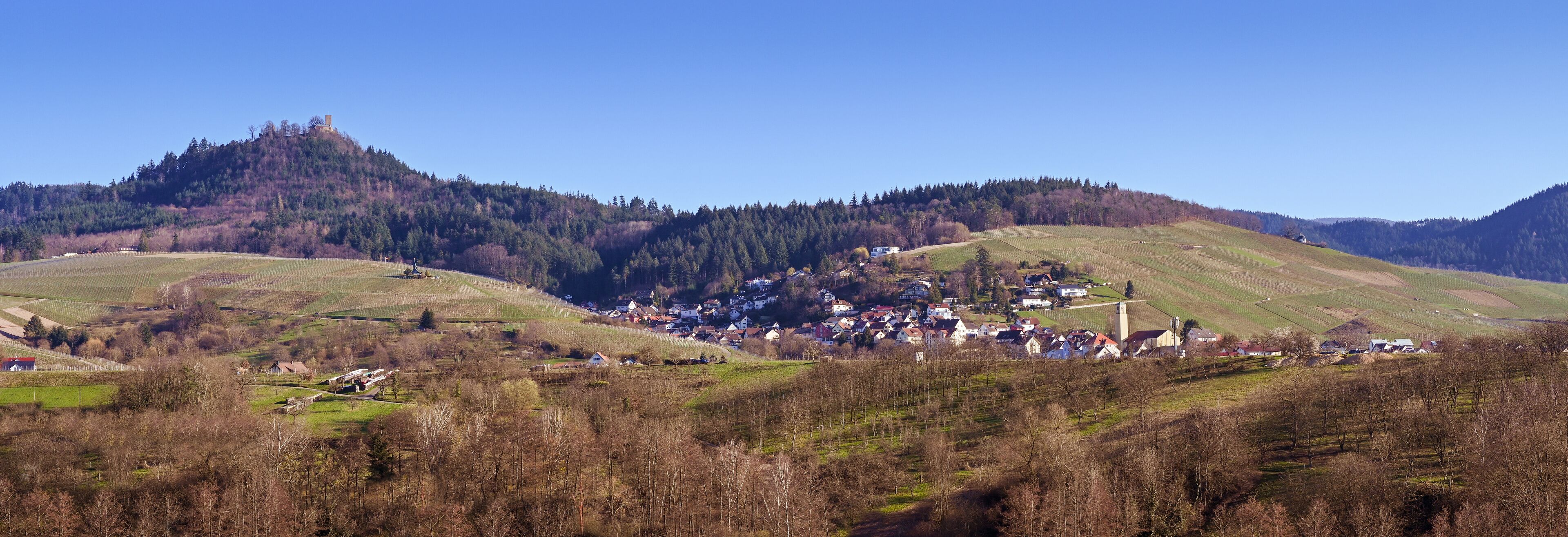 A village nestled in a valley surrounded by hills, with vineyards and a castle on a hill.