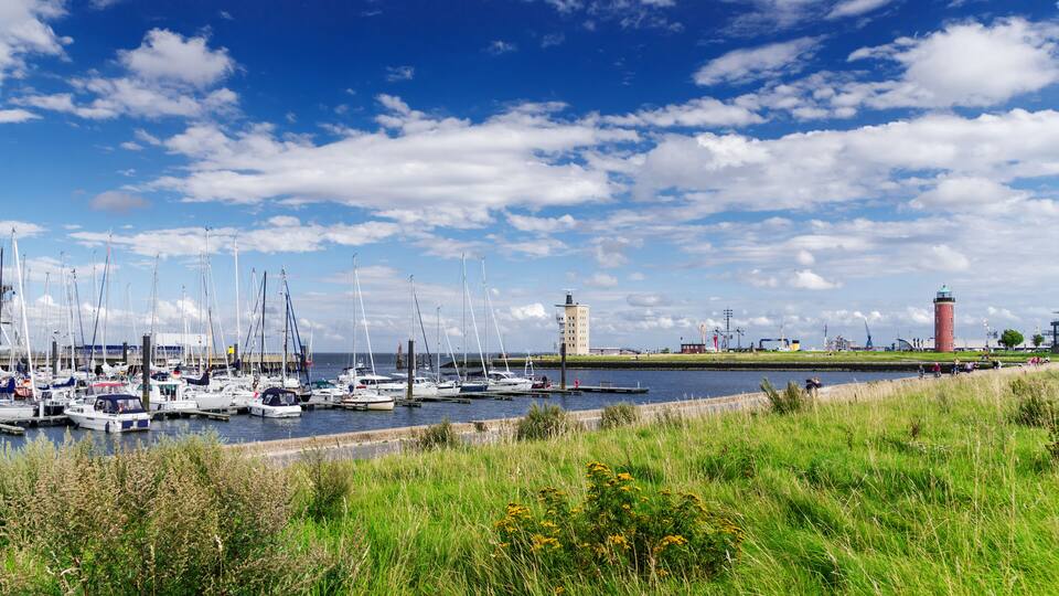 A marina with multiple sailboats docked, a lighthouse, and industrial structures in the background.