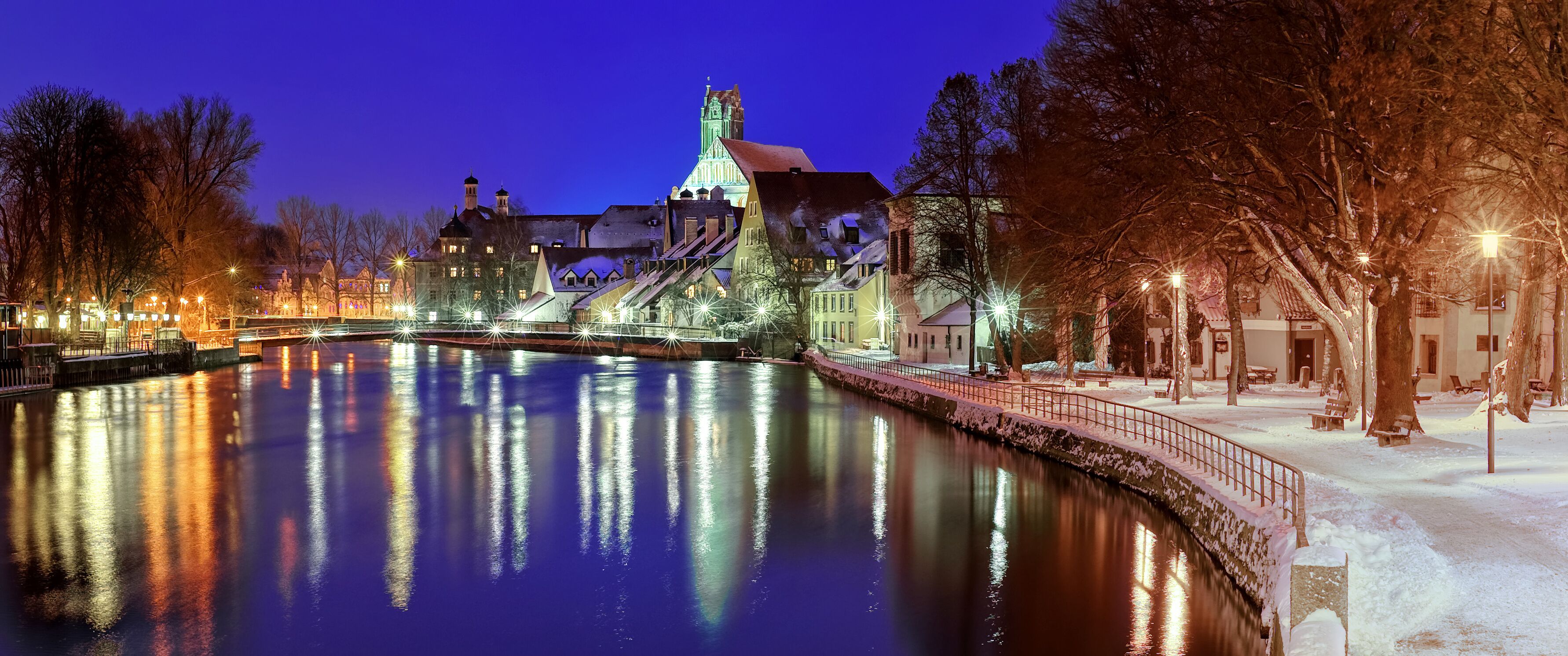 Um rio coberto de neve com edifícios iluminados e uma ponte refletindo luzes.