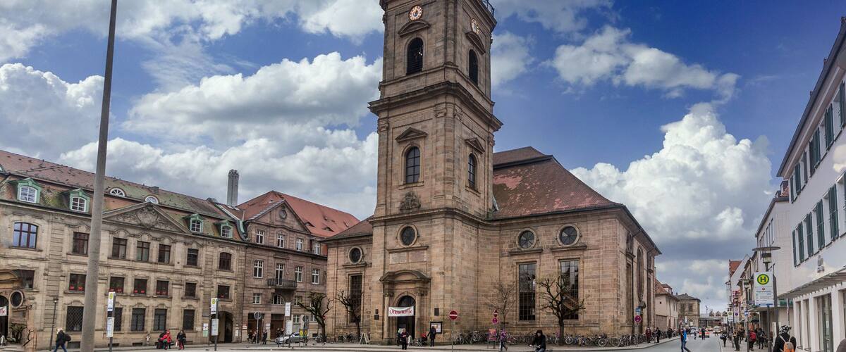A historic building with a prominent clock tower, surrounded by other classic architecture and a clear blue sky with clouds.