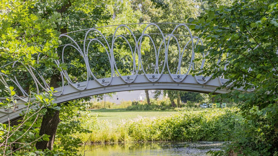 A metal bridge over a calm river, surrounded by lush greenery and trees.