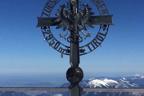 Une croix ornée d’un emblème représentant un aigle et d’une inscription, le tout se détachant sur un paysage montagneux.