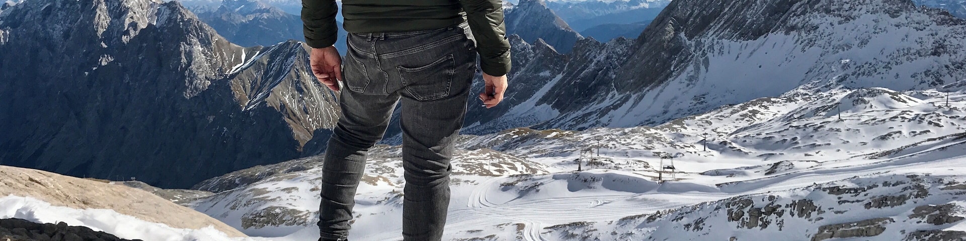 Eine Person steht auf einem felsigen Berggipfel mit Blick auf ein verschneites Tal und entfernte Berge.