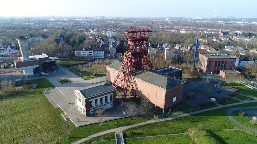 Vista aérea de un paisaje urbano con una imponente torre industrial roja, un gran edificio de ladrillo y un área verde de parque.