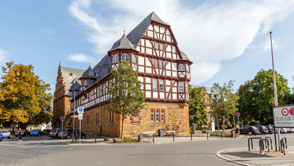 A historic half-timbered building with a stone base and a steeply pitched roof, surrounded by trees and parked cars.