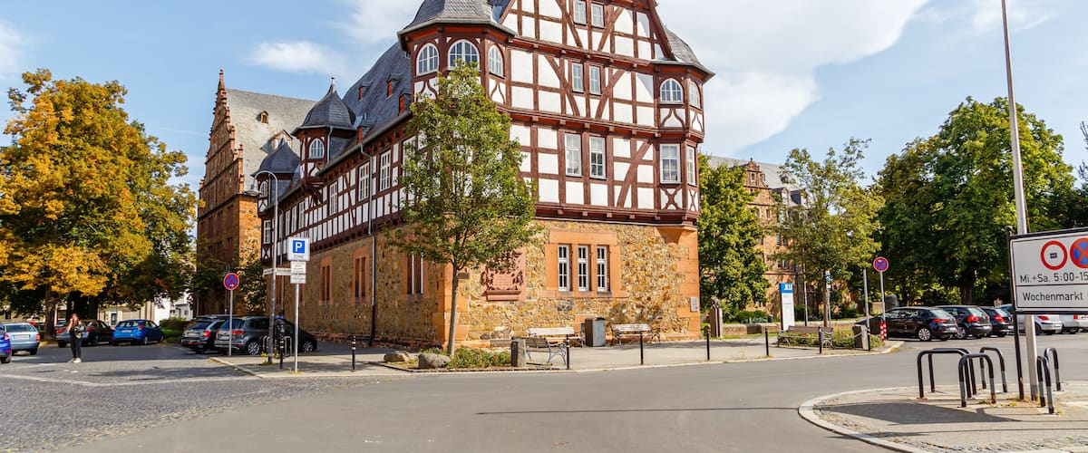 A historic half-timbered building with a stone base and a steeply pitched roof, surrounded by trees and parked cars.