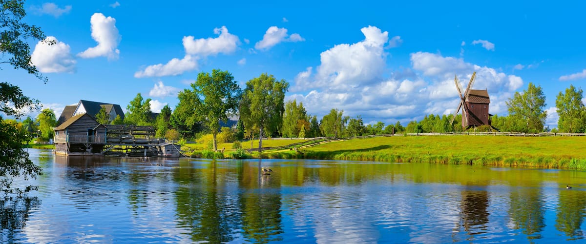 Eine historische Mühle an einem stillen See, umgeben von üppigem Grün und blauem Himmel.