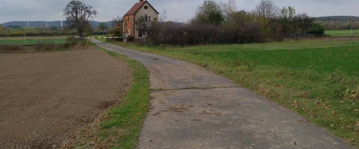 A rural landscape with a dirt path leading to a stone house surrounded by fields and trees.