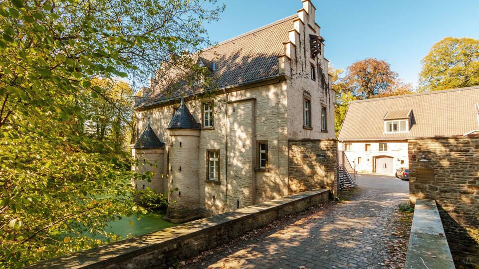 A historic stone building with a turret, a cobblestone pathway, and a tree-lined canal.