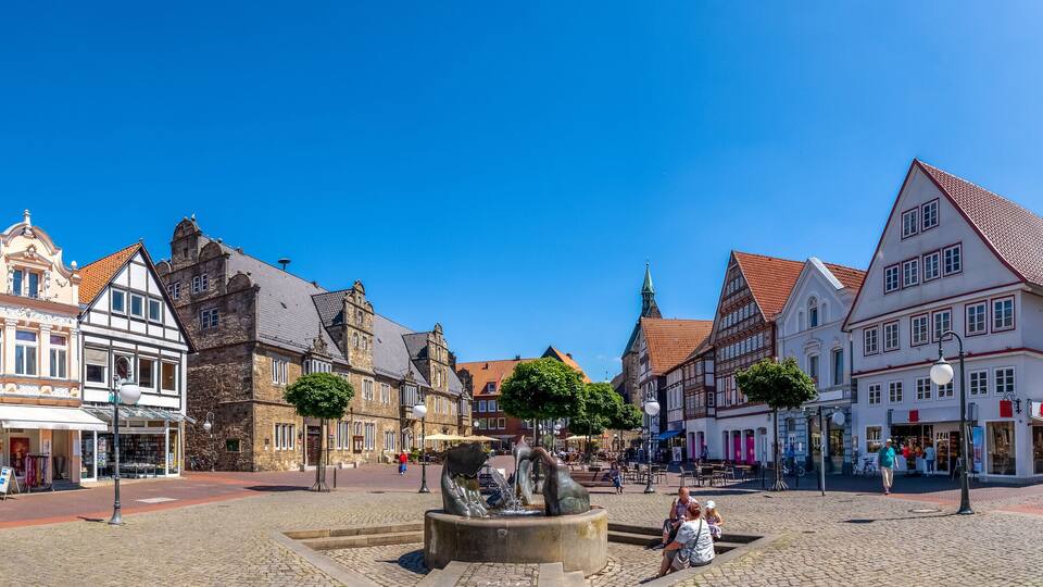 A cobblestone square with historic buildings, a fountain, and a clear blue sky.