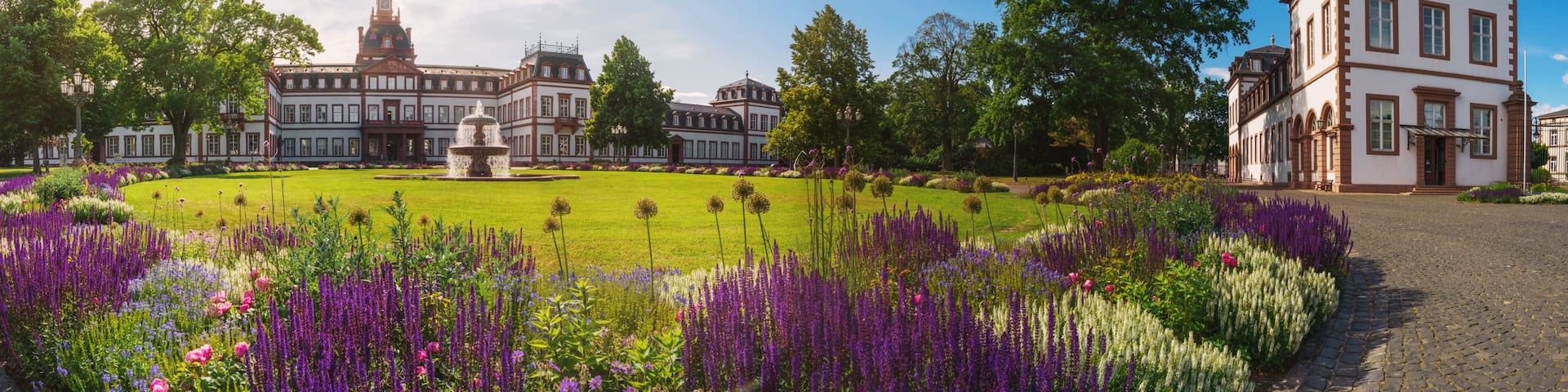 A historic building with a fountain, surrounded by a well-manicured garden with purple flowers.