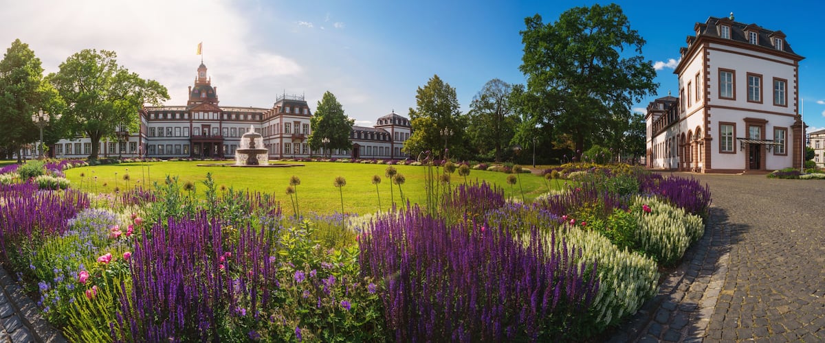 A historic building with a fountain, surrounded by a well-manicured garden with purple flowers.