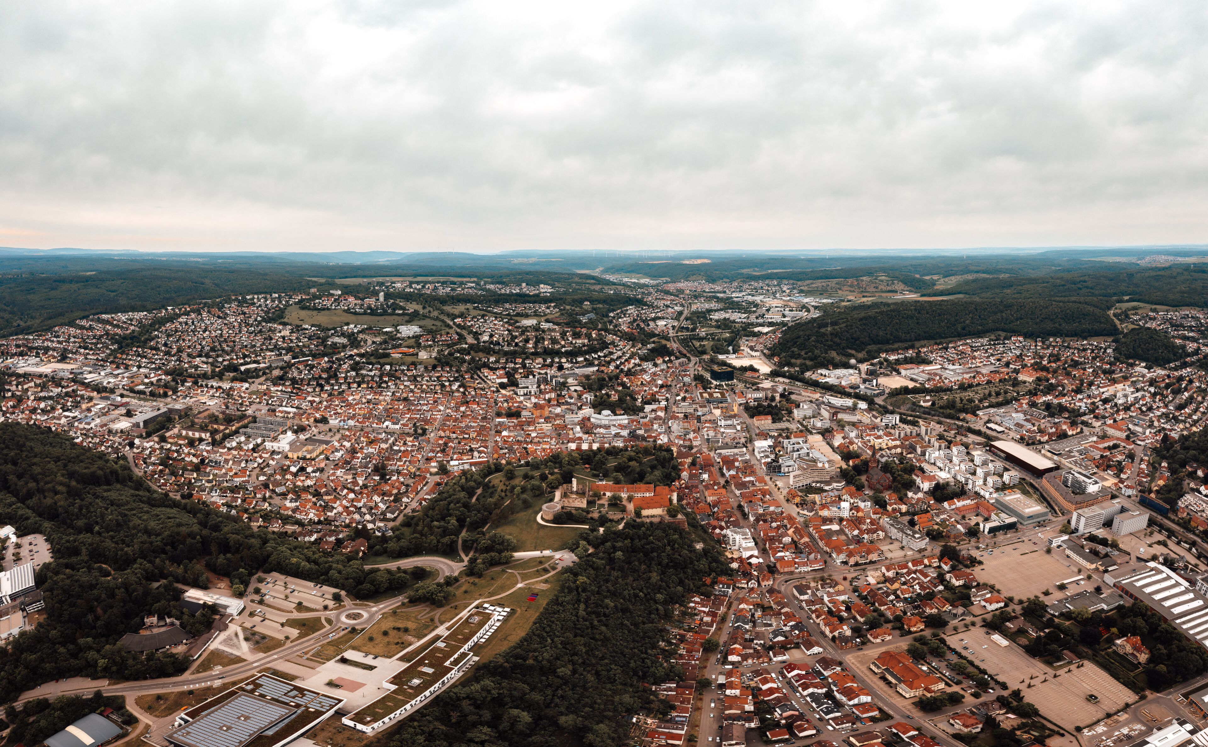 An aerial view of a densely populated city with a mix of residential and commercial buildings, surrounded by green hills and a large open area.