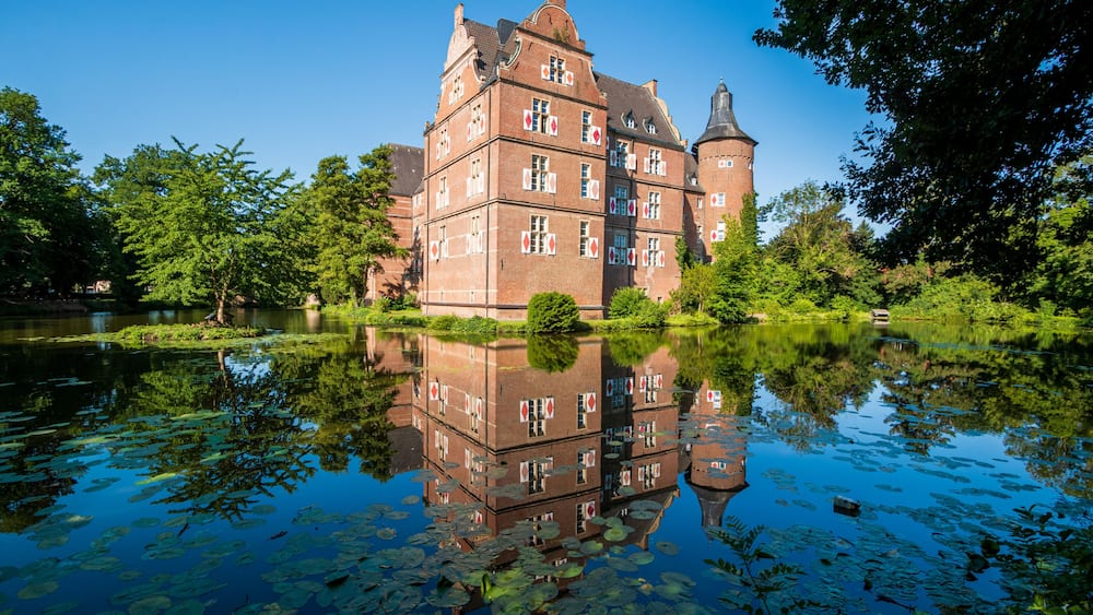 A historic building with a tower, surrounded by a calm lake with lily pads, and lush greenery.