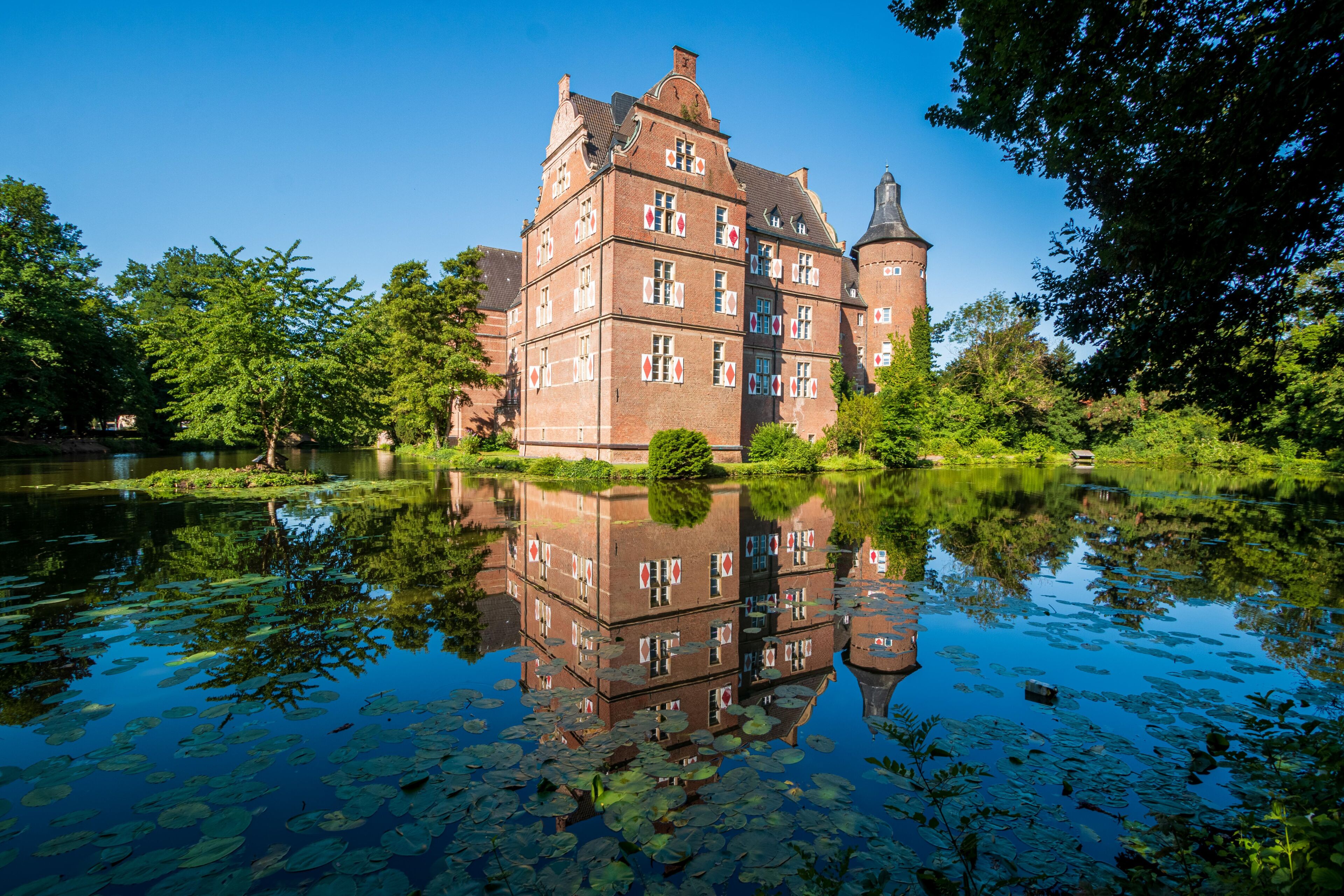 A historic building with a tower, surrounded by a calm lake with lily pads, and lush greenery.
