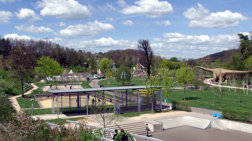 A dinosaur exhibit with a large model dinosaur, a fenced area, and a park with trees and a clear sky.