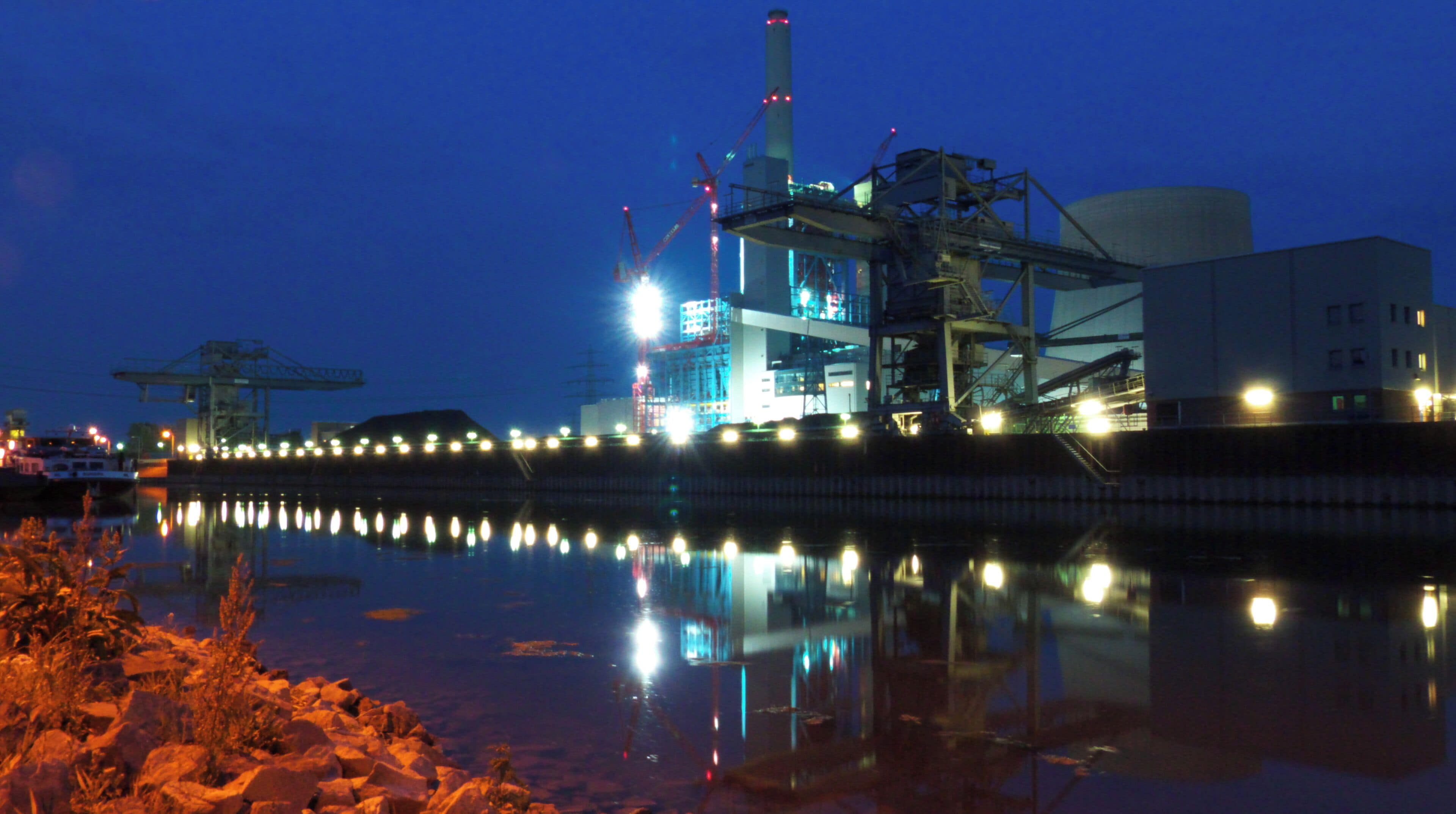 A large industrial facility with cranes and a tall chimney, reflecting on the water.