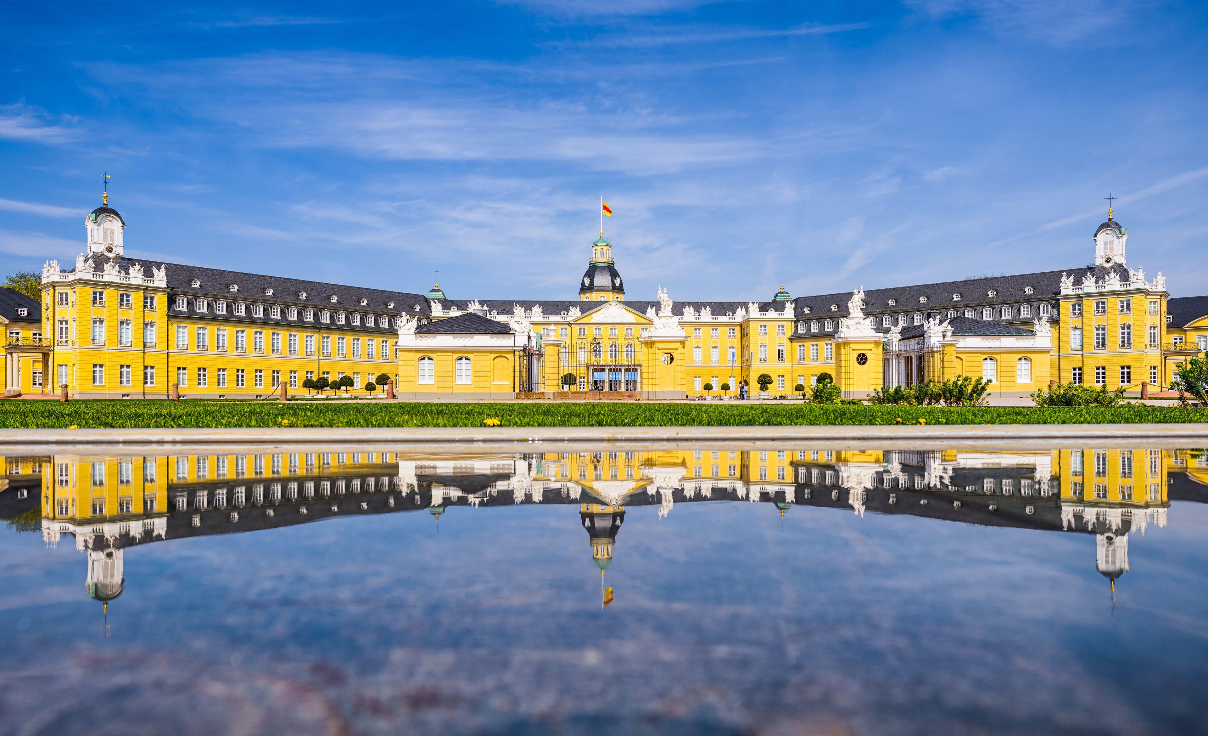 A large yellow building with a reflection in the water, a clear blue sky, and a flag on top.