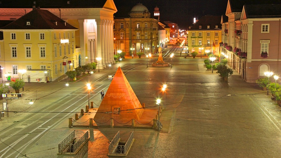 Ein nächtlicher Blick auf einen Stadtplatz mit einem markanten, orangefarbenen Pyramidenmonument, umgeben von historischen Gebäuden mit beleuchteten Fassaden.