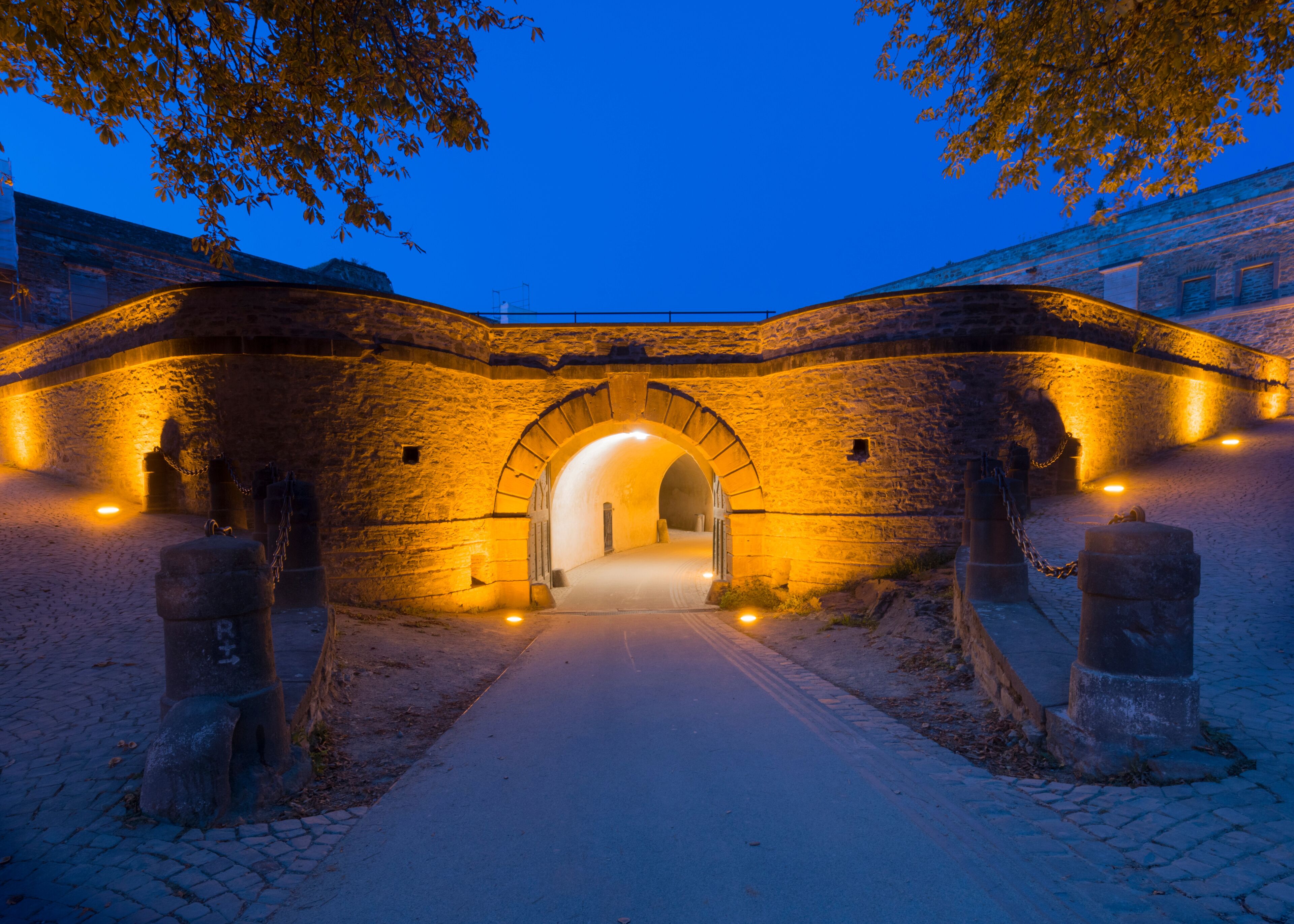 Un arco de piedra iluminado que da paso a un túnel, rodeado de caminos empedrados y pilares de piedra.