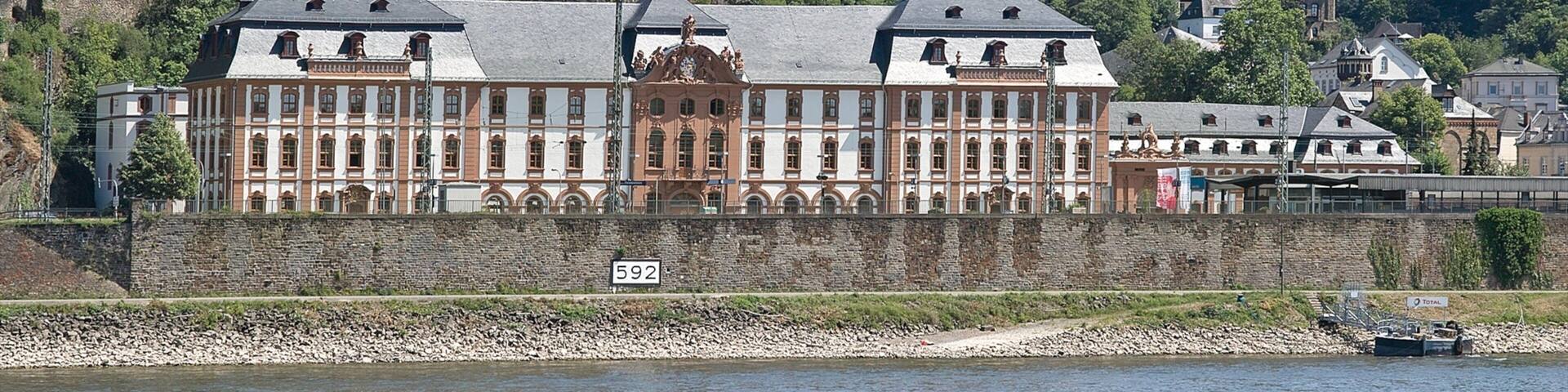 A historic building with a river in the foreground, a cable car system, and a hill with ruins in the background.